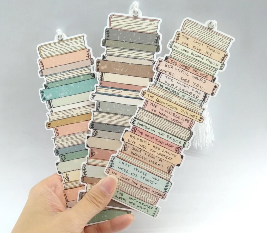 Three bookmarks being held by a white hand. Each one is a stack of books, and the bookmark on the right has book titles written on the spine.