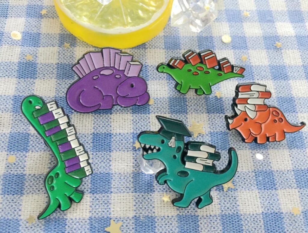 Five brightly colored dinosaur enamel pins, each one featuring books either on their heads, under their chins, or as part of their body. 