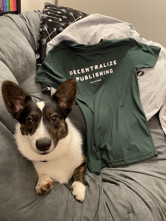 A photo of Gwen, a black and white Cardigan Welsh Corgi, sitting on a gray chair with a green shirt that says, "Decentralize Publishing" in the background.