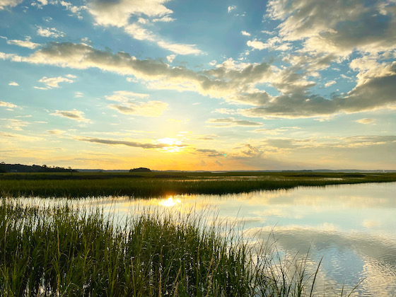 a photo of Hilton Head Island at sunset. The clouds above are reflected in the water. Green marsh grass grows in the shallow water