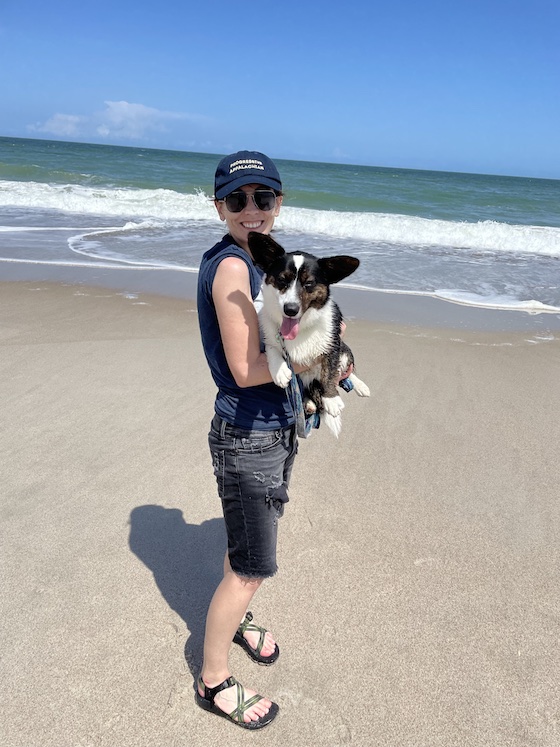 a photo of Kendra, a white woman wear black short, blue t-shirt, blue hat, and chacos. Gwen, a black and white Cardigan Welsh Corgi, sits in Kendra's arms. The ocean can be seen behind them.