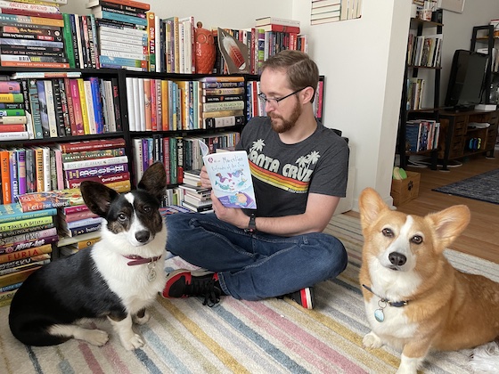 a photo of Sam, a white man with light brown hair and a full beard, reading to Dylan and Gwen. Dylan is a red and white Pembroke Welsh Corgi, and Gwen is a black and white cardigan Welsh corgi. They are all sitting on a multi-colored rug. A bookshelf is behind them.