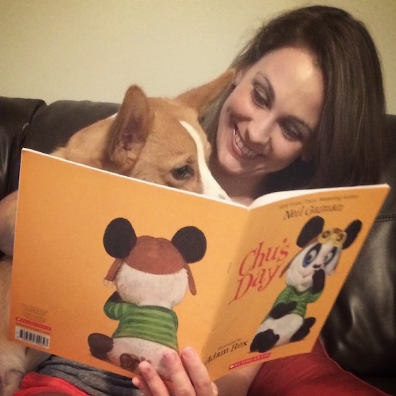 a photo of Kendra, a white woman with brunette hair, holding Dylan, a red and white Pembroke Welsh Corgi. Kendra is reading a children's book to Dylan.