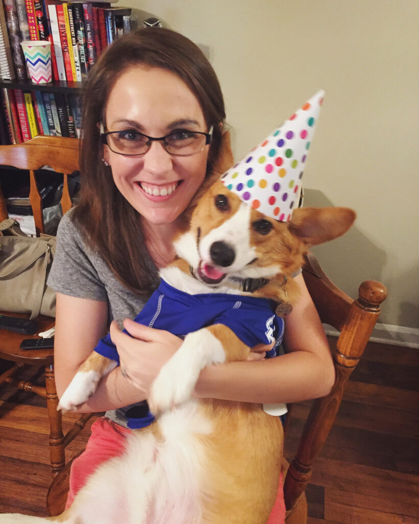 a photo of Dylan, a red and white corgi, being held on Kendra’s lap. Kendra is a white woman with brunette hair. They are both smiling at the camera. Dylan is wearing a birthday hat and a UK wildcats Jersey.