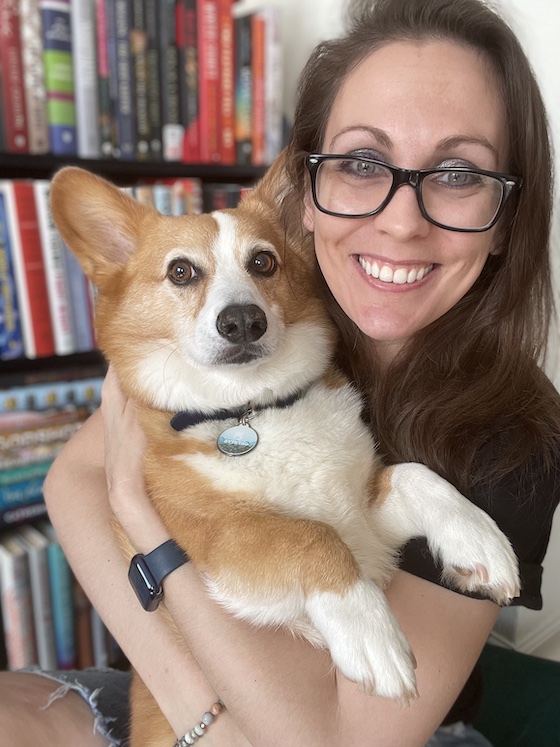 a photo of Kendra, a white woman with brunette hair, holding Dylan, a red and white Pembroke Welsh Corgi. Dylan looks disgruntled, embarrassed even. Kendra is smiling.