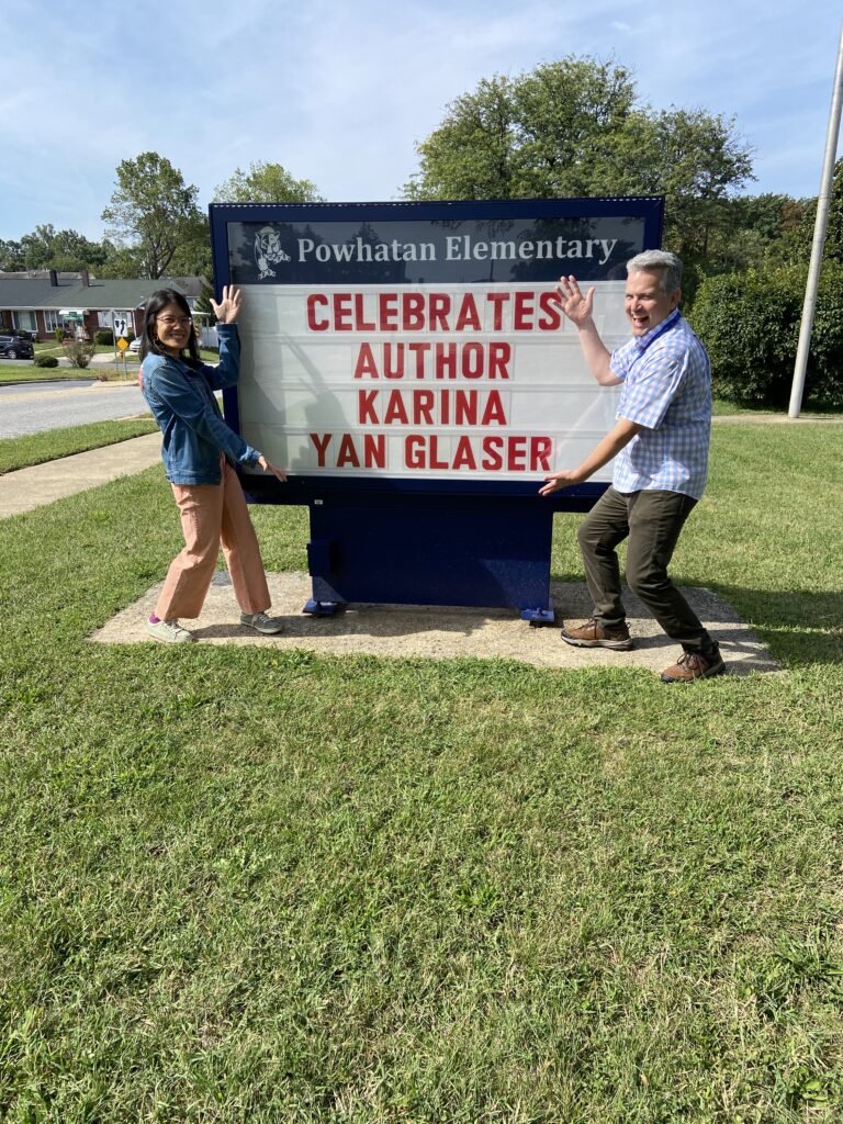 Two people gesturing to a school billboard that says Powhatan Celebrates Author Karina Yan Glaser