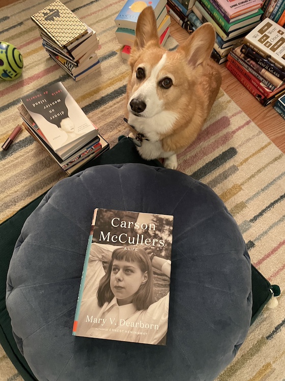 a photo of Dylan, a red and white Pembroke Welsh Corgi, sitting on the carpet surrounded by books. In front of him is a copy of Carson McCullers by Mary V. Dearborn. He's giving the camera a lot of side eye.