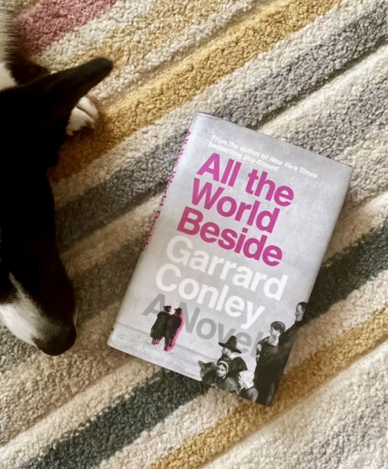 a photo of Gwen, a black and white Cardigan Welsh Corgi, sitting next to a copy of All the World Beside by Garrard Conley